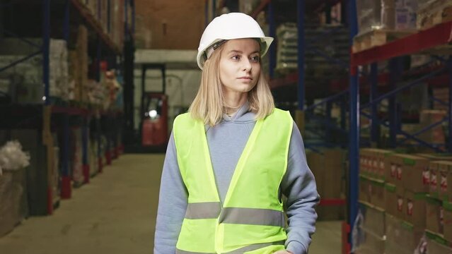 Front view of attractive Caucasian woman wearing safety hardhat and reflective vest standing among shelves rows with goods at large warehouse. Confident supervisor looking at camera.
