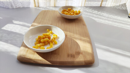 Lot of pieces of canned yellow corn on plate which is on wooden bamboo cutting board on white background. The concept of cooking and delicious healthy food