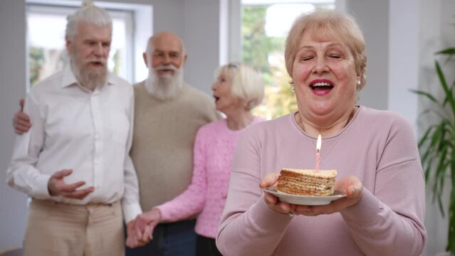 Female retiree smiling blowing out candle making wish on birthday looking back at blurred friends clapping rejoicing. Portrait of Caucasian senior woman celebrating holiday at home indoors