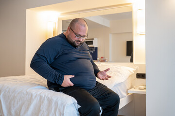 Huge man suffering from extra weight touching stomach sitting on bed in bedroom. Overweight bearded balded middle aged male with unhealthy body noticed problem. Weight control, overeating concept.