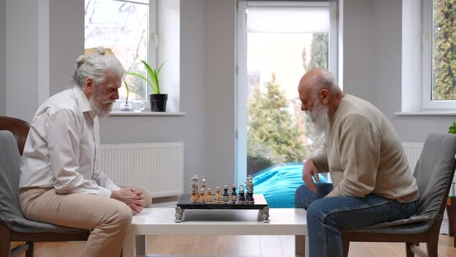 Side view two concentrated old Caucasian men thinking playing chess indoors. Focused male senior friends enjoying game on retirement