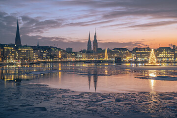 Germany, Hamburg, Ice floating in Alster Lake at dusk with city skyline and glowing Christmas trees in background