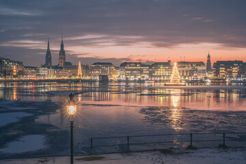 Germany, Hamburg, Ice floating in Alster Lake at dusk with city skyline and glowing Christmas trees in background