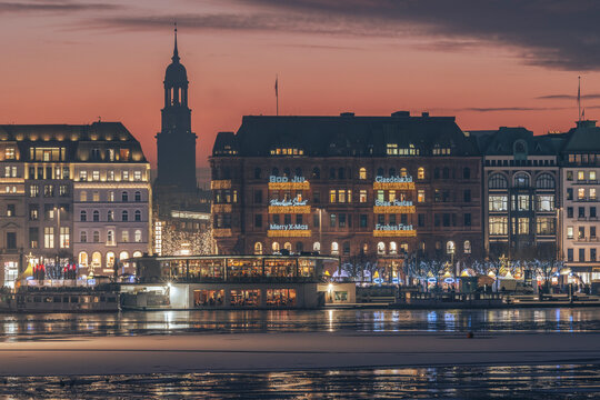 Germany, Hamburg, Christmas decorations hanging on city buildings at dusk with Alster Lake in foreground