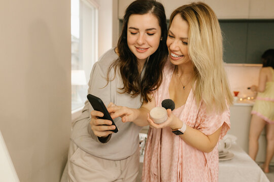 Happy woman sharing smart phone with friend holding beauty product at home