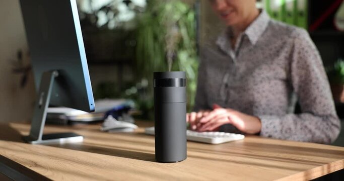 Modern air humidifier device on office desktop. Woman enjoying aromatherapy steam from essential oil diffuser