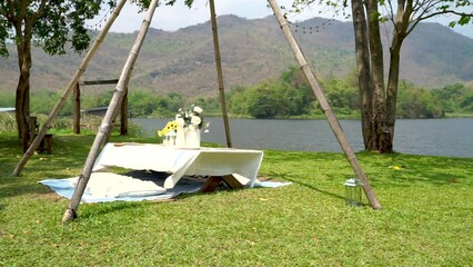 A small, low table with a white cover is decorated with floral arrangements  all set atop a verdant green grass field. The tranquil lake and hills, along with a gentle br