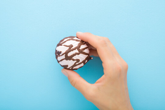 Young adult woman hand holding pink soft zephyr covered with dark brown chocolate glaze on light blue table background. Pastel color. Closeup. Sweet snack. Top down view.