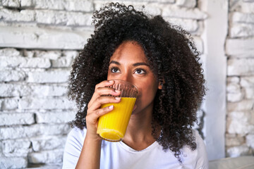 African-American woman enjoys freshly squeezed orange juice on sofa, morning light. - obrazy, fototapety, plakaty