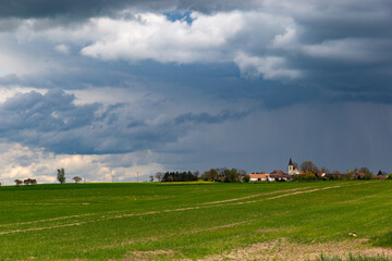 Obraz premium A small European village with a church on a hill, among spring fields, under a dramatic cloudy sky.
