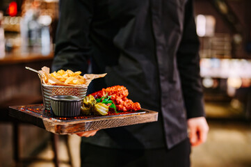 Men's waiter hands hold a tray with a crispy fried breaded chicken breast strips, french fries and sauce on plate in pub