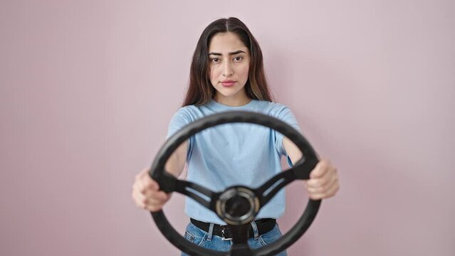 Young beautiful hispanic woman using steering wheel as a driver looking upset over isolated pink background