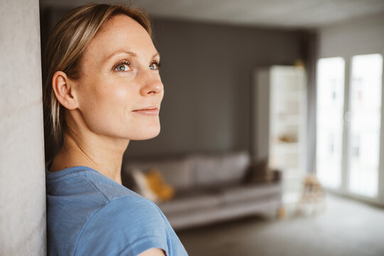 Relaxed blonde woman in blue t-shirt leaning against a pillar in her apartment and looking to the side