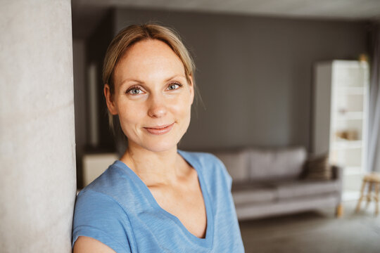 Relaxed blonde woman in blue t-shirt leaning against a pillar in her apartment and looking to the camera