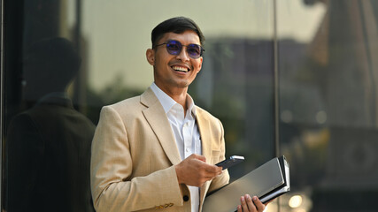 Cheerful asian businessman in photochromic glasses standing in front of modern office building...