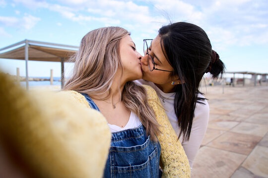 Happy LGBT young lesbian Caucasian couple on tourist vacation kissing for selfie with cell phone outdoors. Gay people in love and romantic attitude. Generation z and photos on social networks. 