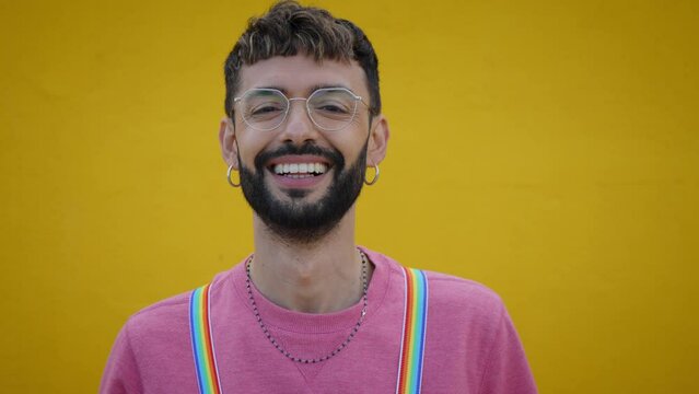 Close up smiling cheerful young Caucasian man with rainbow lgbt suspenders. Gay people posing for video yellow background. Cheerful gay bearded person happy studio isolated. Millennials generation.