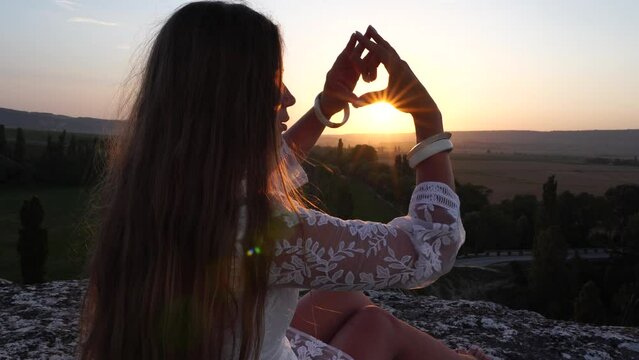 Happy woman in white boho dress making heart sign with hands on sunset in mountains. Romantic woman with long hair standing with her back on the sunset in nature in summer with open hands.