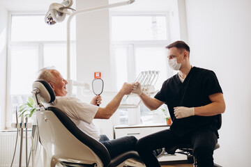 Senior man sitting at dentist chair holding saliva ejector while doctor fixing his tooth.