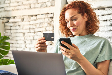 Fototapeta premium Young redhair woman sitting on her sofa doing online shopping on laptop in morning light
