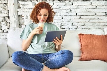 .Redhead woman on sofa, sipping milk, watching videos on tablet for a cozy morning.