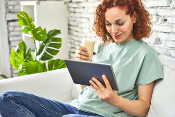 .Redhead woman on sofa, sipping milk, watching videos on tablet for a cozy morning.