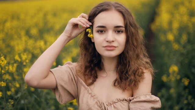 An attractive girl puts a yellow flower behind her ear and smiles. Female portrait in nature on the background of a blooming rapeseed field. Slow motion