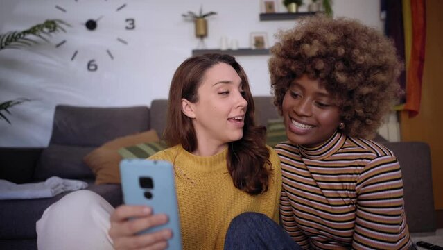 Front view of Multiracial lesbian couple using smartphone while spending time at home in the living room, laughing with social media videos and online shopping. LGBTQ pride month.