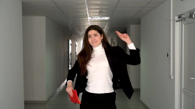Enthusiastic young brunette in smart clothing with red notebook dancing expressively in the middle of empty grey lit hallway of office building 