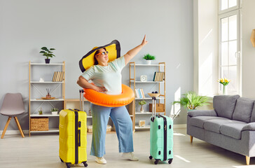 Funny woman going on summer holiday. Portrait of happy fat woman tourist in casual clothes, big sun hat, sunglasses and beach ring standing in living room with two vacation travel suitcases