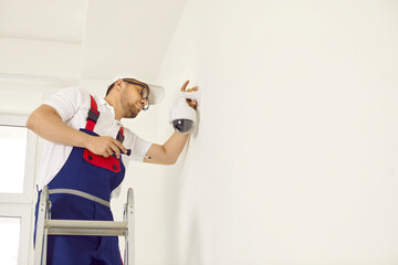 Male worker installing a security camera on a white wall at home. Young man in a work uniform uses...