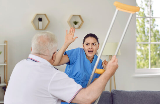 Angry elderly man threatening nurse to hit her with crutch. Enraged exasperated senior man abusing and threatening scared female caretaker, volunteer with his cane