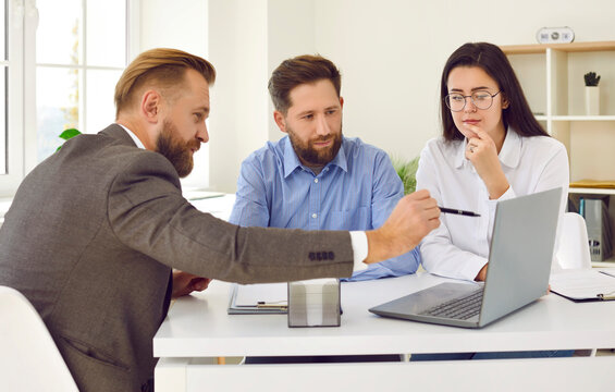 Young couple meeting with professional realtor. Real estate agent sitting at office table, talking to clients, offering options, pointing at laptop computer. Mortgage loan, property purchase concept