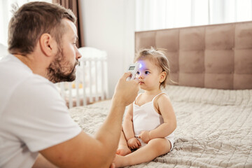 An ill little girl is sitting on the bed while her father s measuring temperature with thermometer.