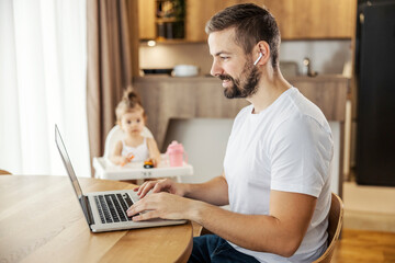 A happy casual businessman is sitting at home and working on a laptop while his daughter playing in blurry background.