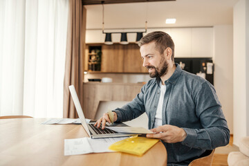 A happy man is paying bills online on a laptop. He is managing home finances.