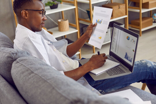 Young business man doing research. African American man sitting on comfortable couch at home, working on modern laptop computer, studying financial report, looking at digital slides, and taking notes
