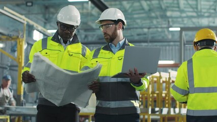 Two men move and communicate with each other on construction site. Engineers dressed in yellow vests and helmets. African American architect holding blueprints. Worker is gesturing and holding laptop. - Powered by Adobe