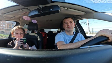 A 40-year-old Caucasian father and his 12-year-old son drive together in a car, with the sun shining and palm trees visible outside the window. The son is absorbed in his smartphone, periodically
