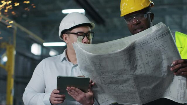 Asian engineer holding tablet and writing something down. African-American man in yellow vest and hard hat asks for advice, holds blueprint. Workers in special clothes cooperate at construction site.