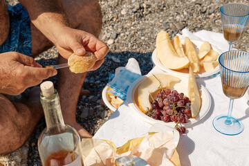 Summer beach picnic. Hands of man preparing appetizers for picnic with fresh fruit, bread, cheese and wine outdoors at seaside at the beach.