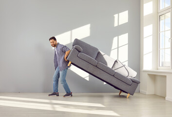 Man moving the sofa at home. Full shot of a happy young man moving his gray couch while moving the...