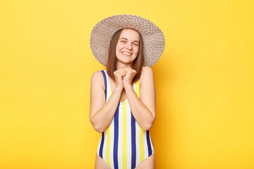Woman with cheerful expression wearing striped swimsuit and sun hat while standing isolated on yellow background, looking directly at the camera, enjoying peaceful day at beach.