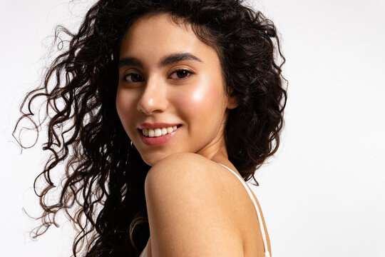 Young woman with thick black and curly hair and glowing skin smiles and looks at the camera..