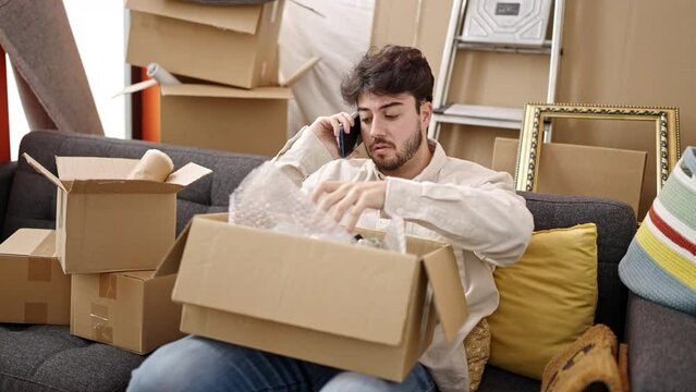 Young hispanic man speaking on the phone unpacking cardboard box at new home