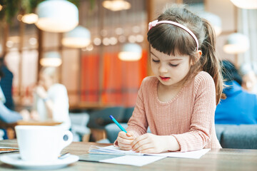 Fototapeta na wymiar Cute little girl writing something in a notebook sitting in a cafe