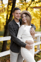 Happy stylish pregnant woman and man hugs, kiss, touches belly and listen baby's kicks, walk outdoor in autumn park. Pregnancy, motherhood and parenthood concept. Family is expecting for unborn baby.