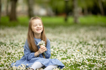 Happy little girl in a cotton dress sitting in a field of daisies in the summer at sunset and laughing.