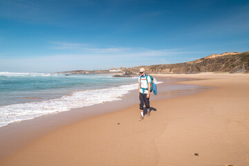 Backpacker walks along Praia do Almograve with a smile on his face. The joy of moving and discovering new places. Odemira region, western Portugal. Wandering along the Fisherman Trail, Rota Vicentina
