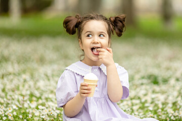 Fototapeta premium Funny happy toddler girl wearing cotton summer dress sitting on a green lawn eating vanilla and chocolate ice cream cone in a sunny garden or park. Space for text